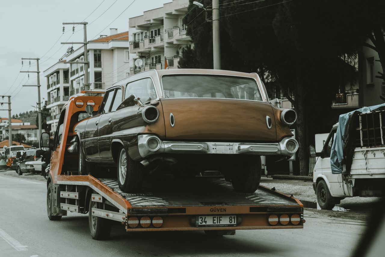 hero-img-01 A vintage car loaded on a tow truck in a city street, surrounded by modern buildings.