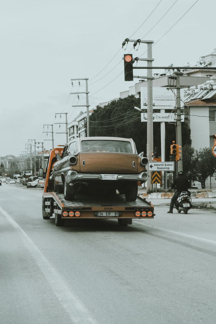 hero-img-02 A vintage car is being towed through city streets on a tow truck, showcasing an urban environment.